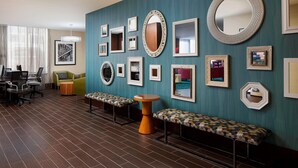 Un hall d’hôtel moderne avec un mur de couleur turquoise, plusieurs miroirs encadrés et un espace salon comprenant une table ronde orange et des bancs à motifs.
