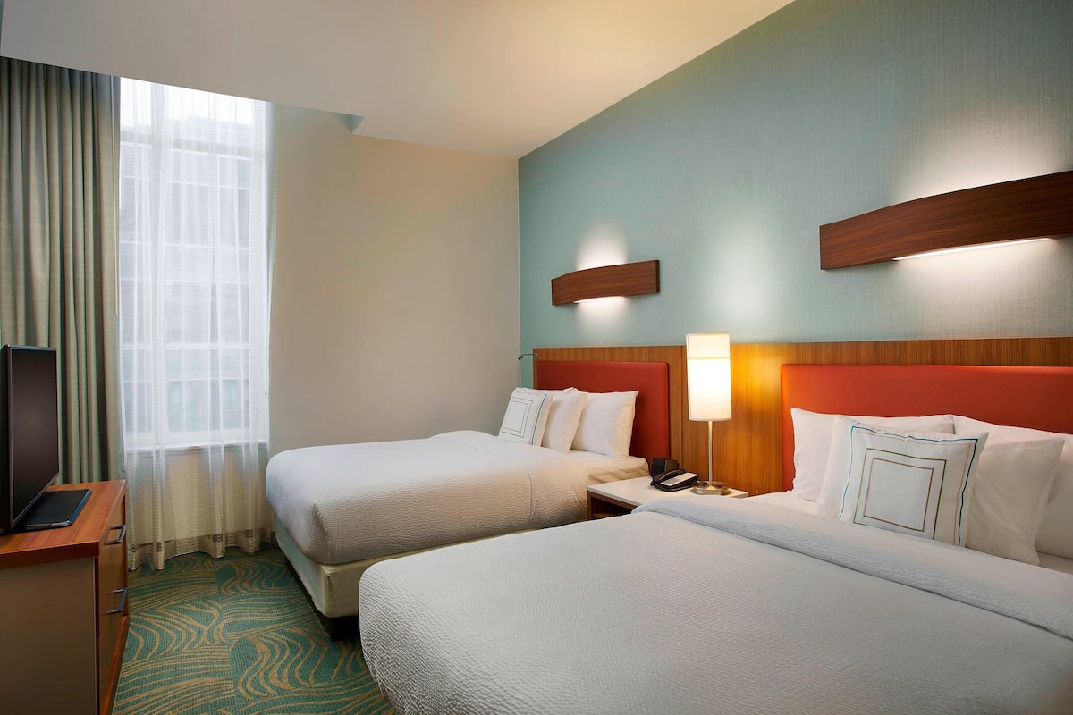 A hotel room with two beds, a desk with a laptop, a bedside table with a lamp, and a window with sheer curtains.
