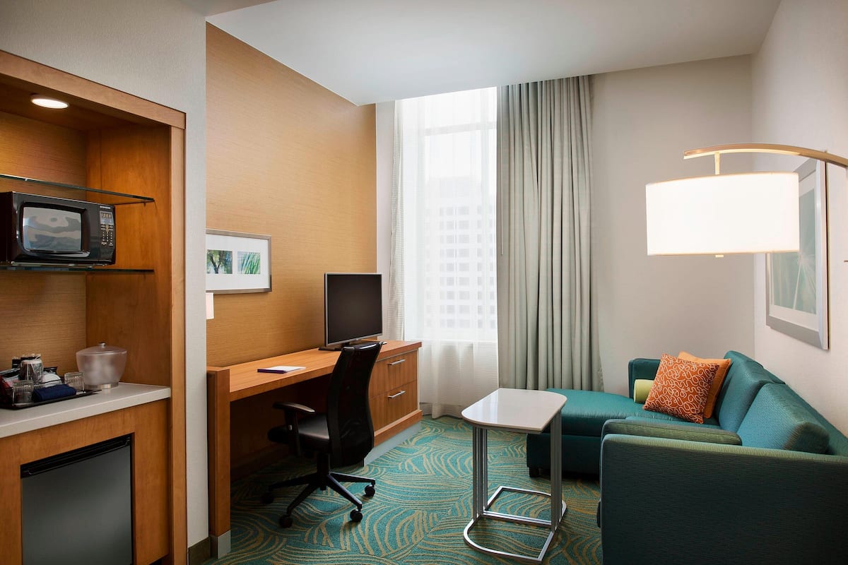 A hotel room with a desk, a small kitchenette, a sofa, and a large window with curtains.