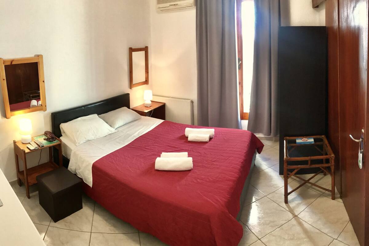 A hotel room with a large bed, a bedside table with a lamp, a TV mounted on the wall, and a small wooden stool.