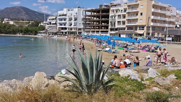 A beach with people sunbathing and swimming, surrounded by buildings and a mountain in the background. - Atlantis Hotel (Agios Nikolaos)