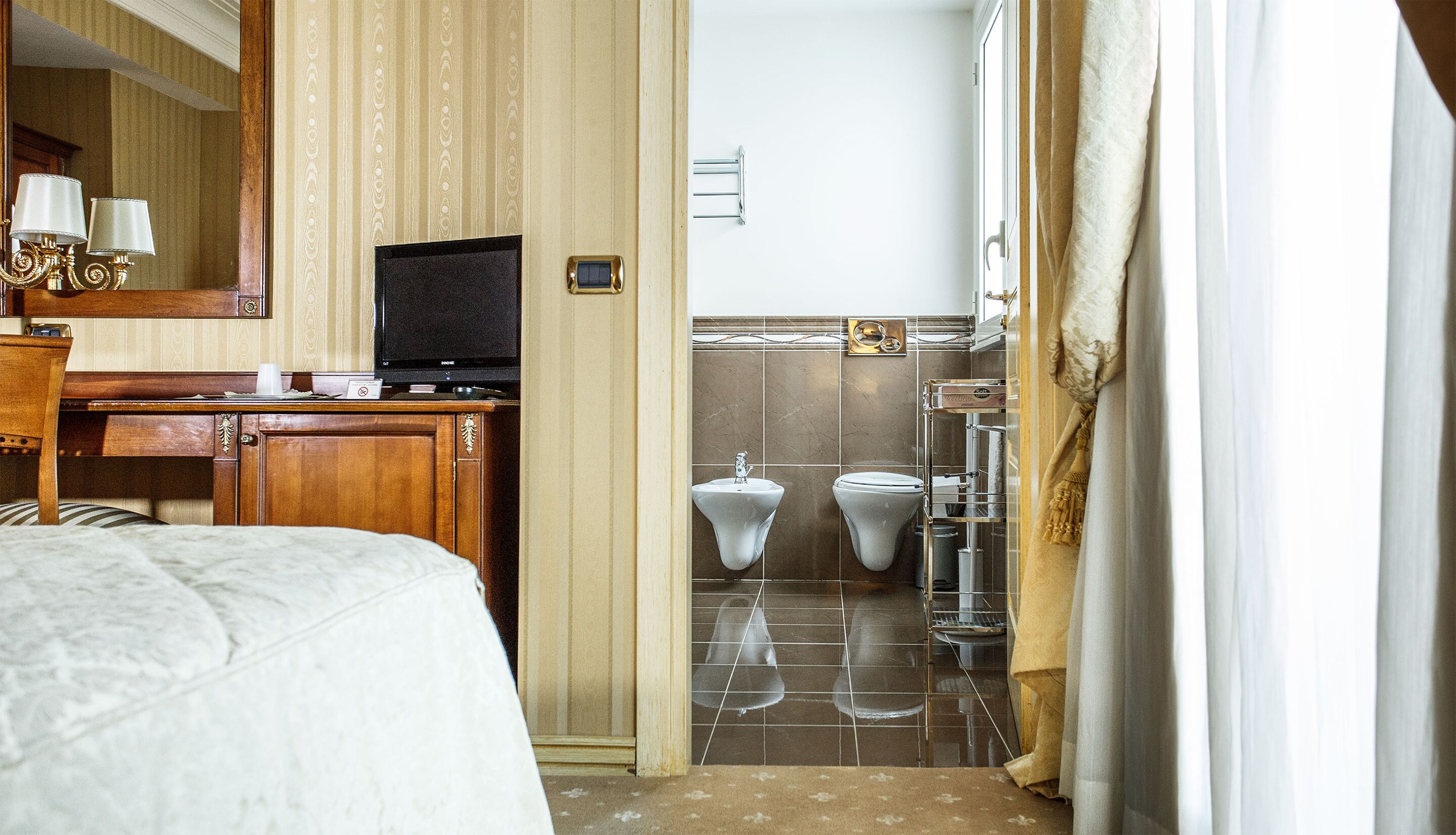 Habitación de hotel con una cama, un escritorio con televisor, y un baño con dos inodoros y un lavamanos.