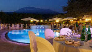 Un área de comedor al aire libre con piscina, mesas dispuestas para una comida y sombrillas que brindan sombra.