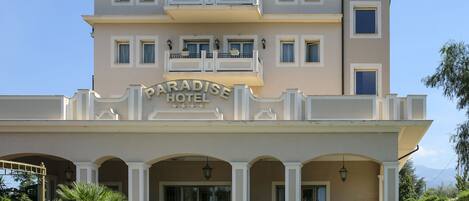 A multi-story hotel with a prominent "Paradise Hotel" sign, a balcony with railings, and a well-maintained front garden with palm trees and spherical garden lights.