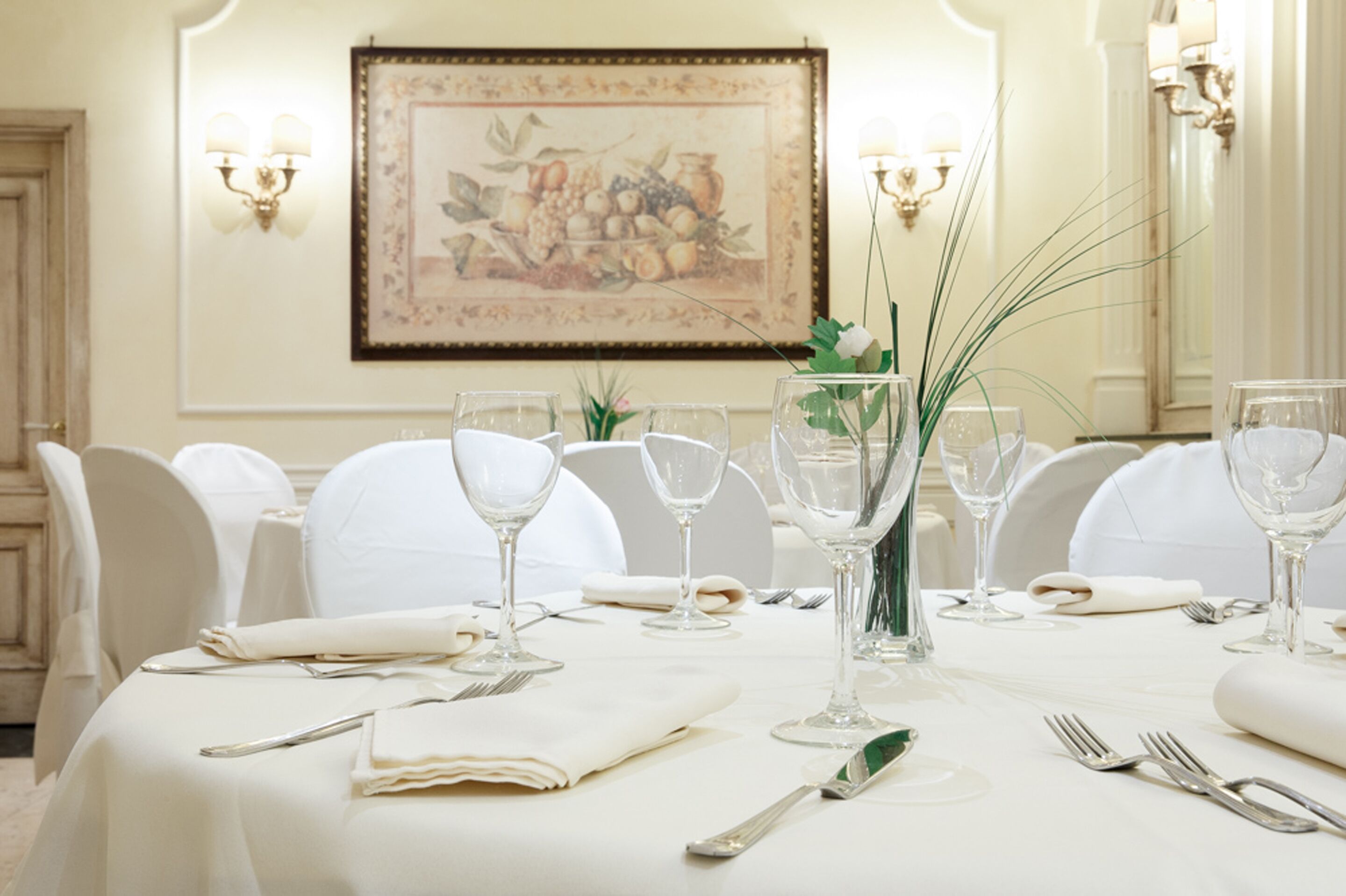 A dining table set with white tablecloth, glassware, and silverware, with a framed artwork on the wall behind.