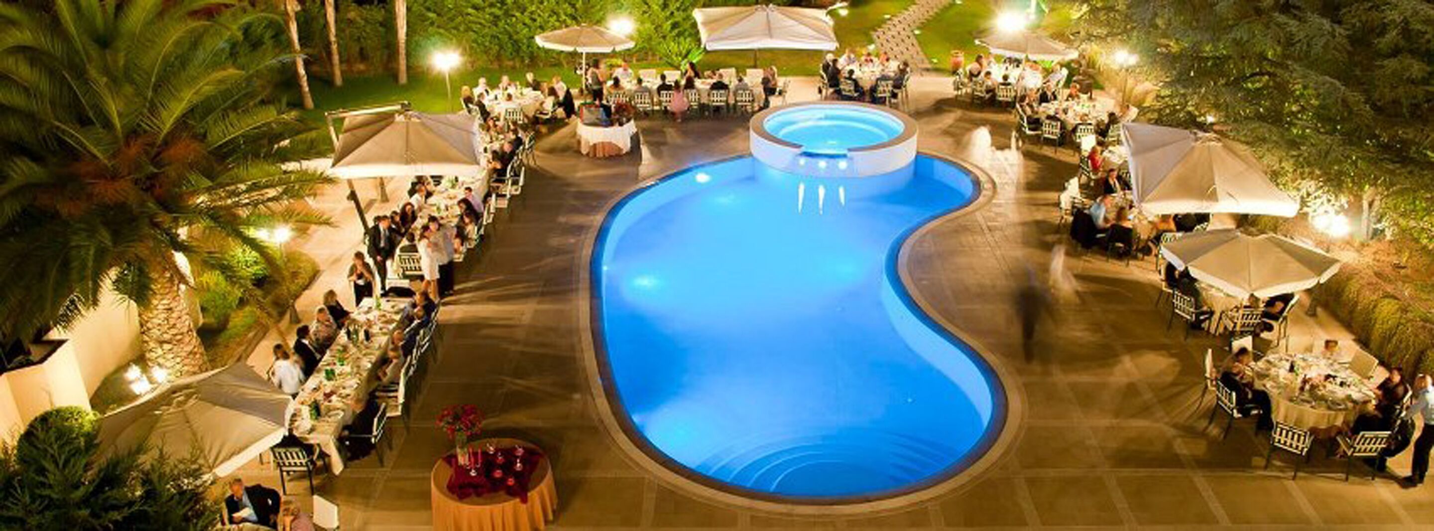 An outdoor pool area with tables and umbrellas, surrounded by palm trees.