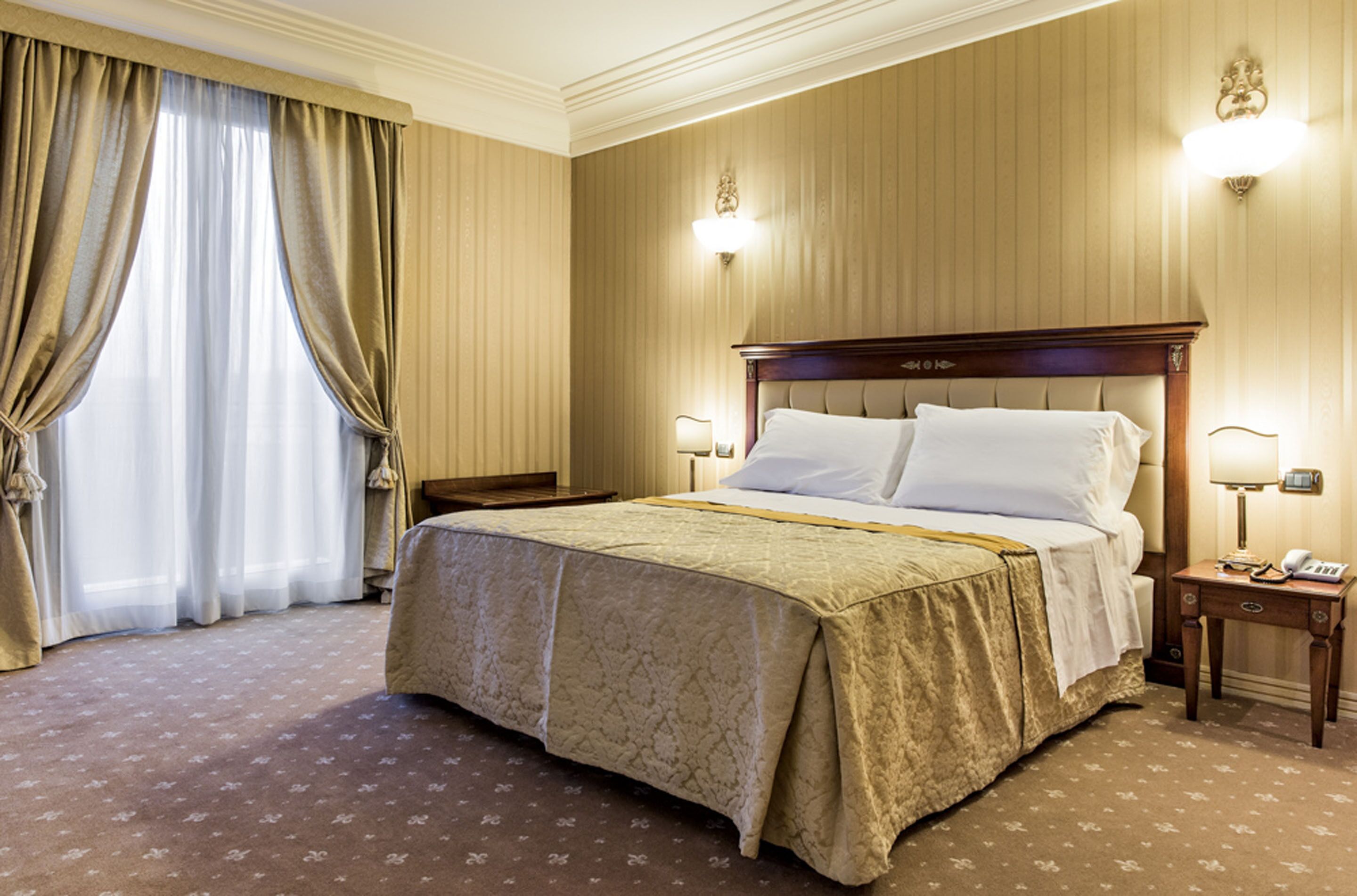 Habitación de hotel con una cama grande, una mesita de noche, una lámpara y una ventana con cortinas.