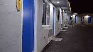 Um corredor de hotel com portas e janelas azuis, chão de concreto e escada que leva a um andar superior.