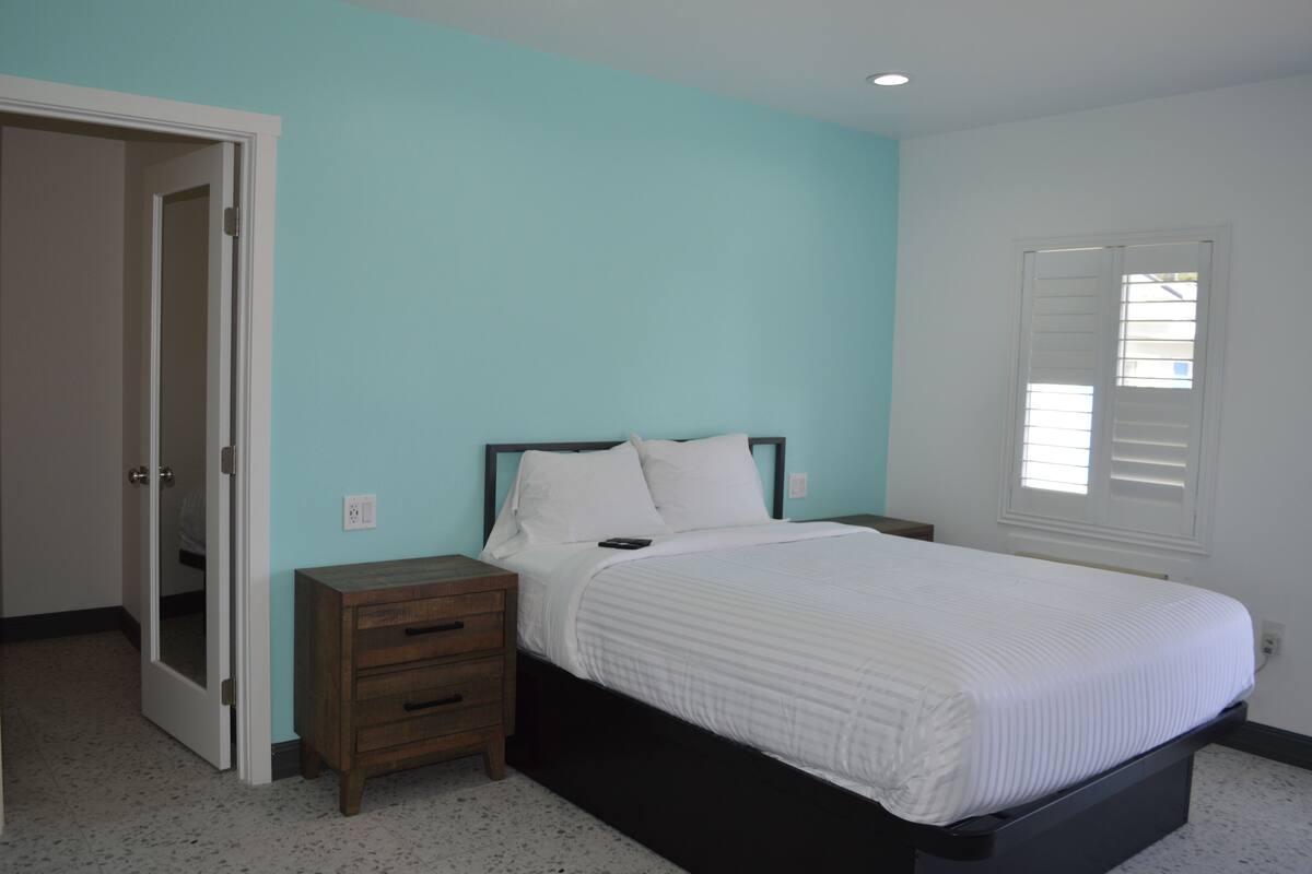 A bedroom with a bed, a nightstand, a wooden dresser, a door, and a window with blinds.