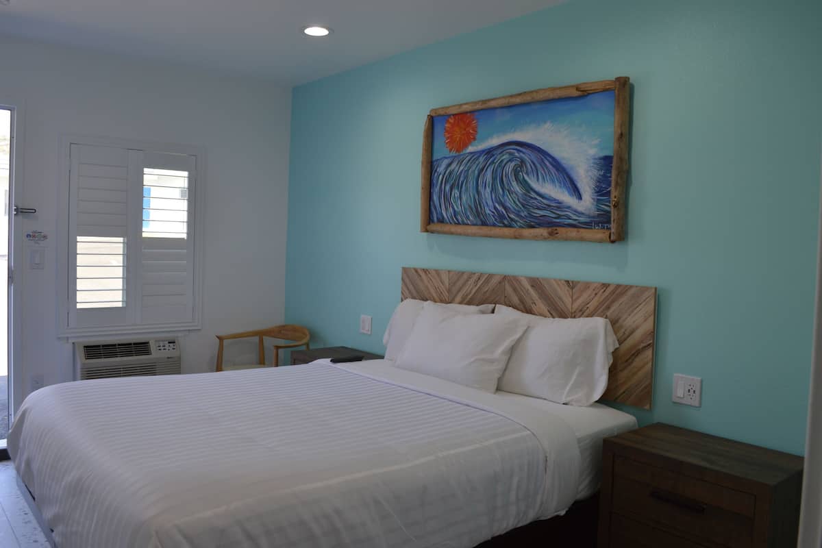 A bedroom with a bed, a nightstand, a wooden headboard, a painting of a wave, and a window with blinds.