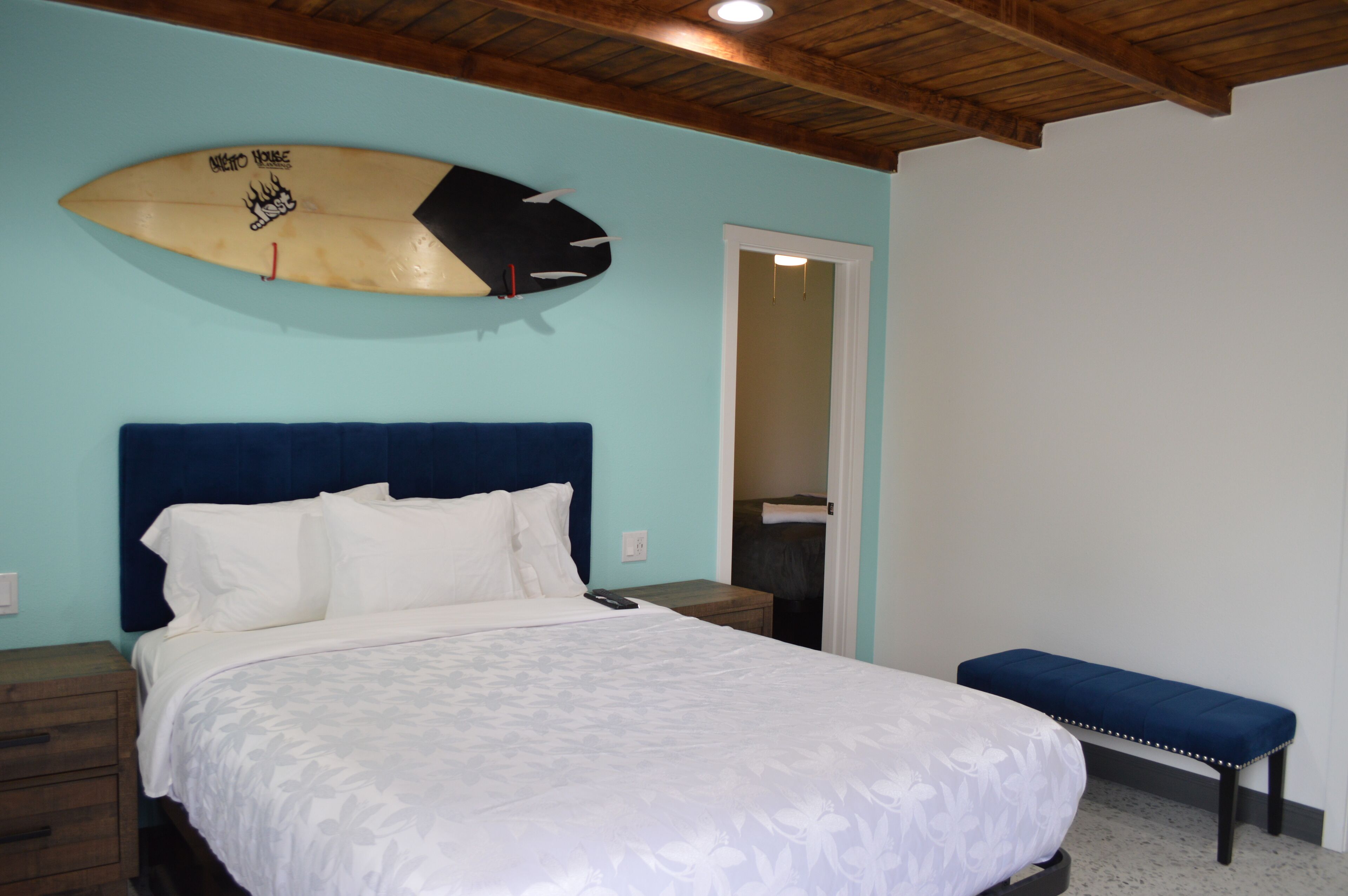 Un dormitorio con cama, cabecera azul, banqueta y una tabla de surf en la pared.