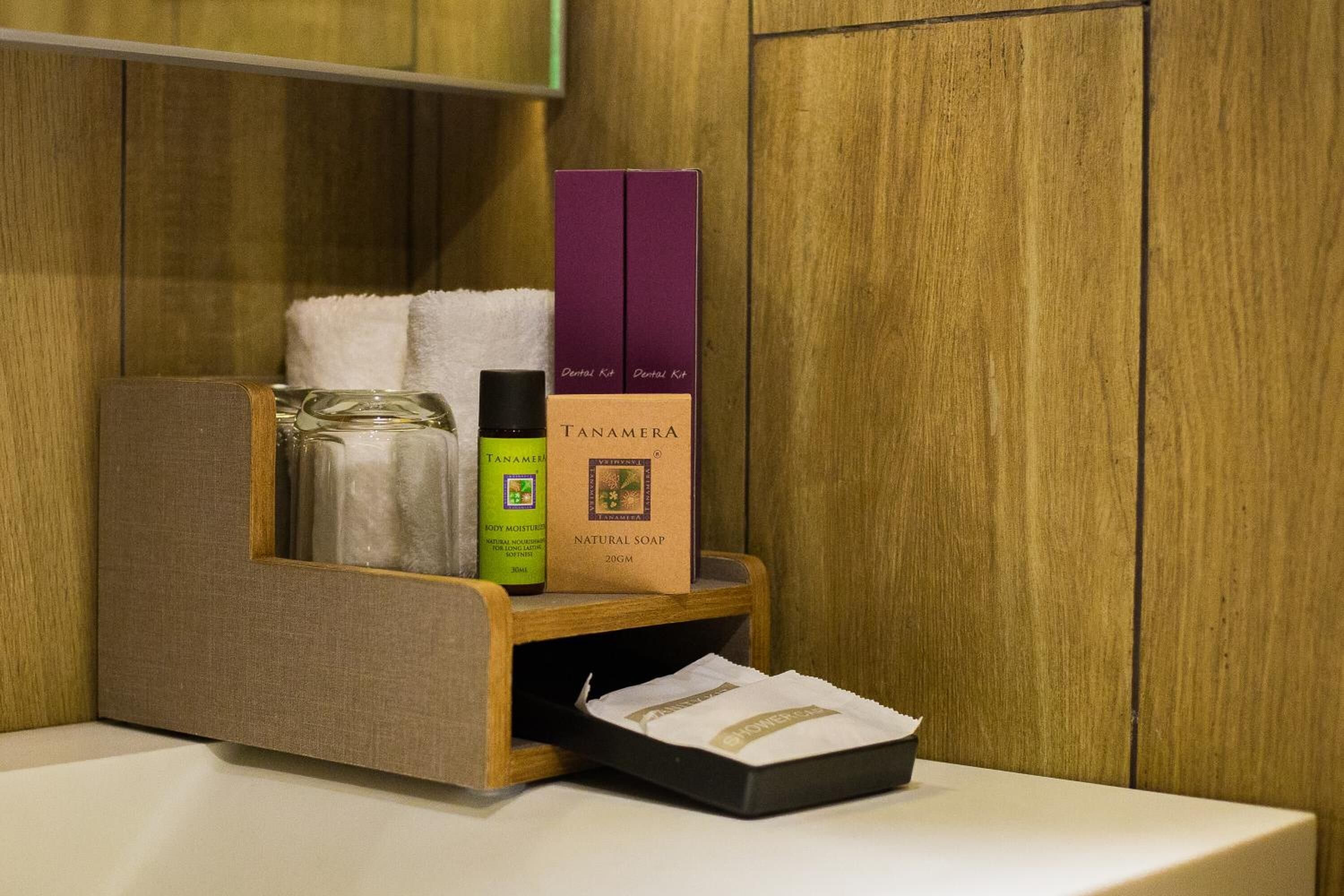 A wooden shelf with a bottle of Tanamera body moisturizer, a box of Tanamera natural soap, and a purple box labeled "Dental Kit" in a hotel setting.