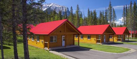 A row of log cabins with red roofs, set in a forested area with a mountain in the background.