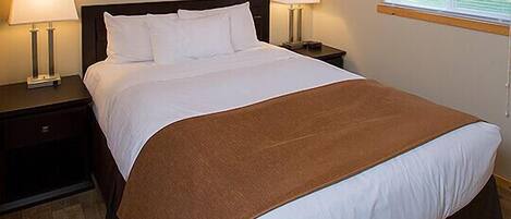 A neatly made bed with a brown bedspread, two bedside lamps, a framed picture on the wall, and a window with blinds.