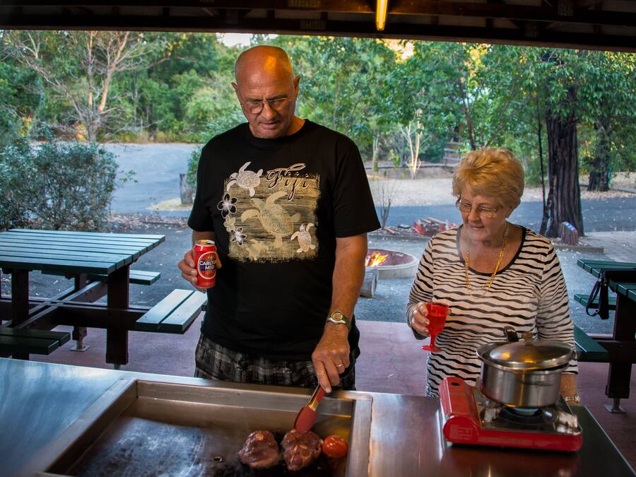 Um homem e uma mulher cozinham ao ar livre em uma estação de churrasco, com uma churrasqueira, um braseiro e mesas de piquenique ao fundo.