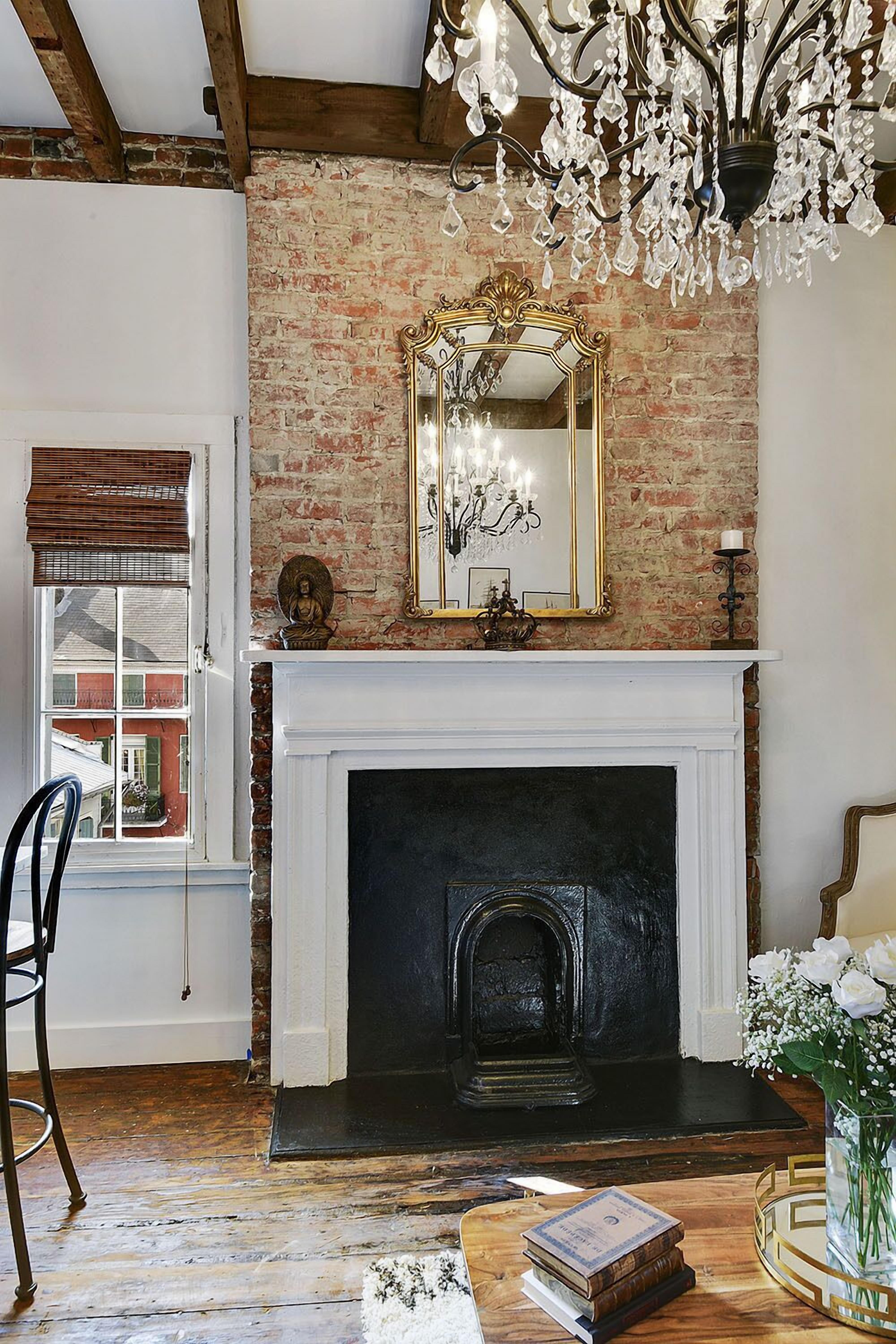 A room with a brick wall, a white fireplace, a chandelier, and a mirror above the fireplace.