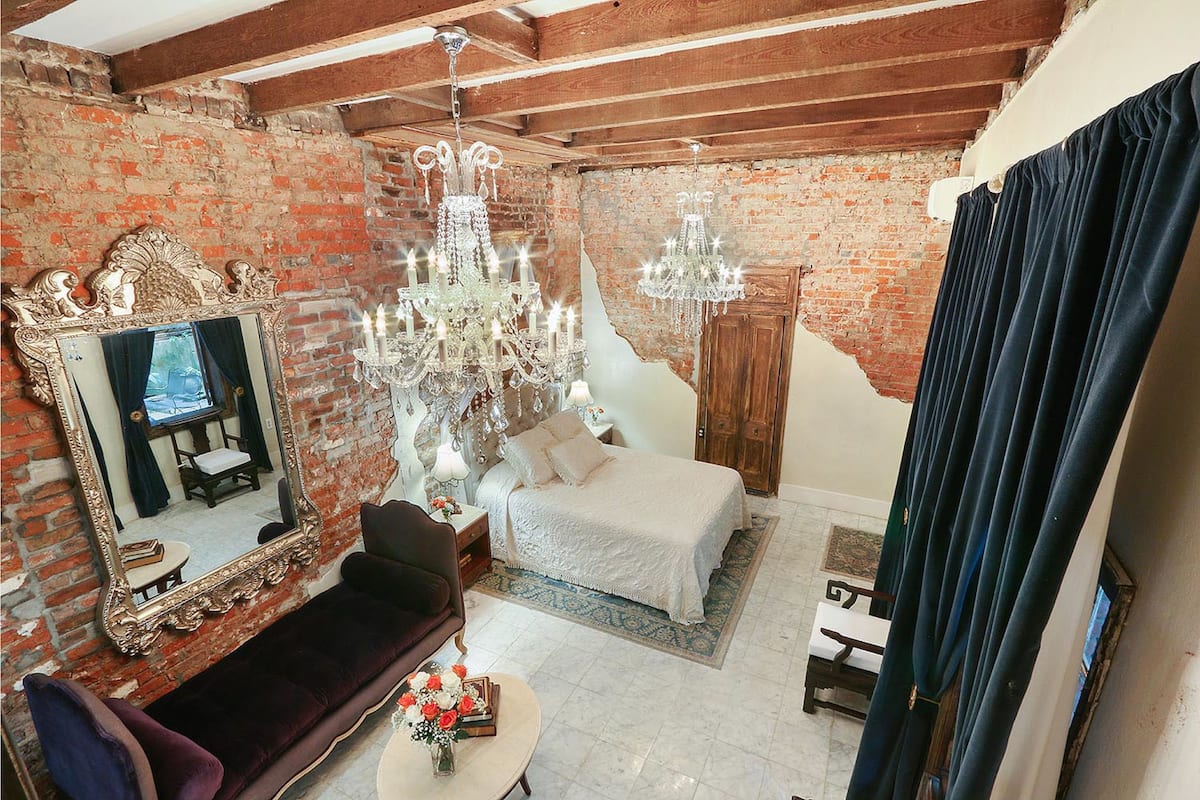 A room with exposed brick walls, a large ornate mirror, a chandelier, a bed with white linens, and a dark blue curtain.