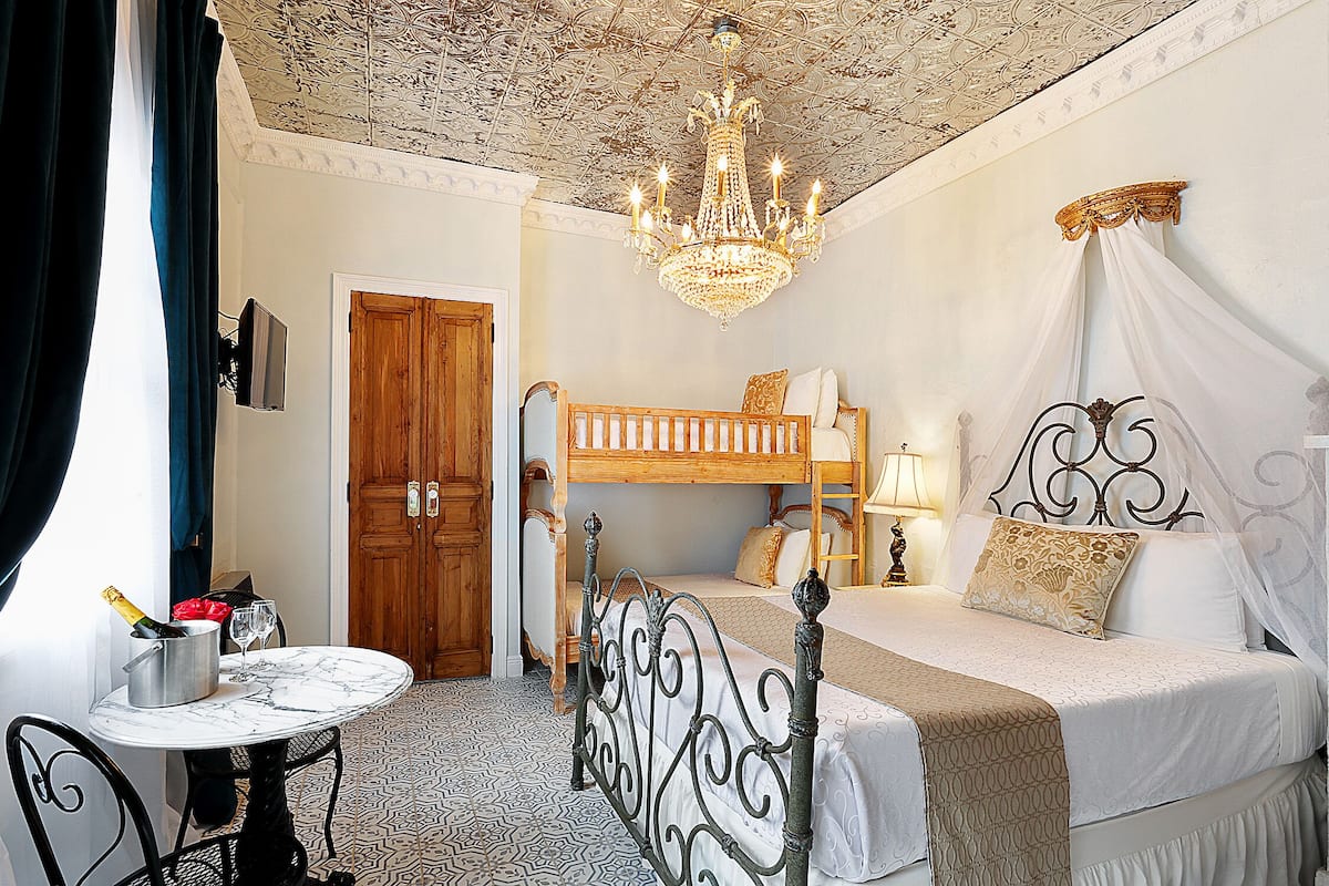A room with a bunk bed, a chandelier, a table with a bottle and glasses, and a patterned floor.