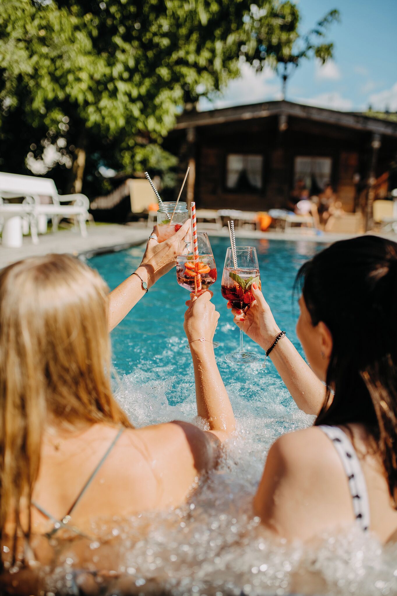 Dos personas brindando con bebidas en un área de piscina con un edificio de madera al fondo.
