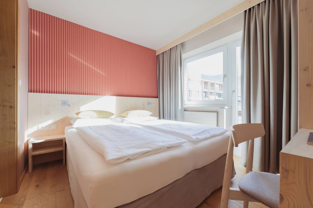 Una habitación de hotel con cama, escritorio, silla y ventana con cortinas.