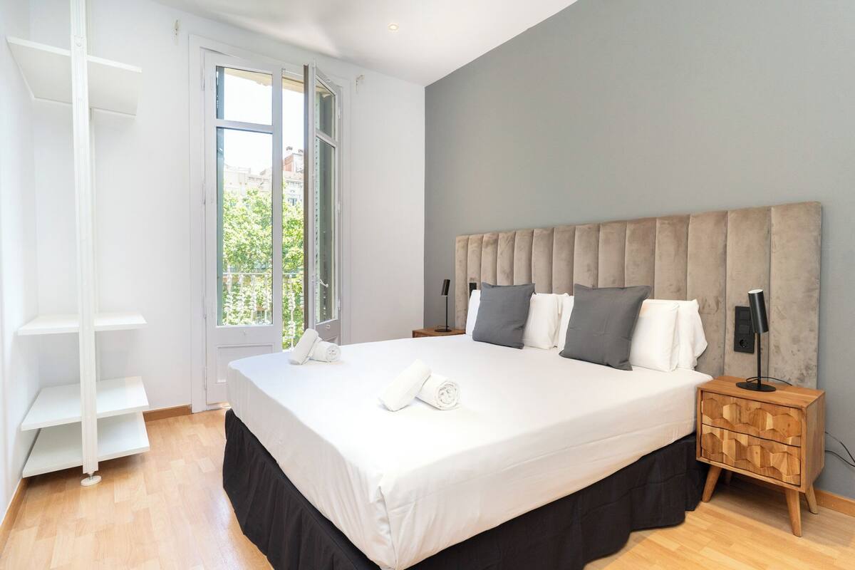 A modern bedroom with a large bed, a wooden nightstand, a bedside table, and a window with a view of greenery.