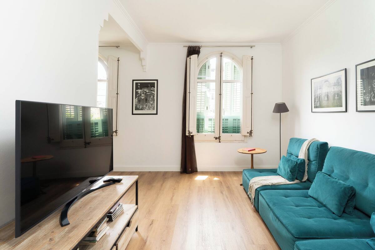 A modern living room with a flat-screen TV, teal sofa, wooden flooring, and a large window with curtains.