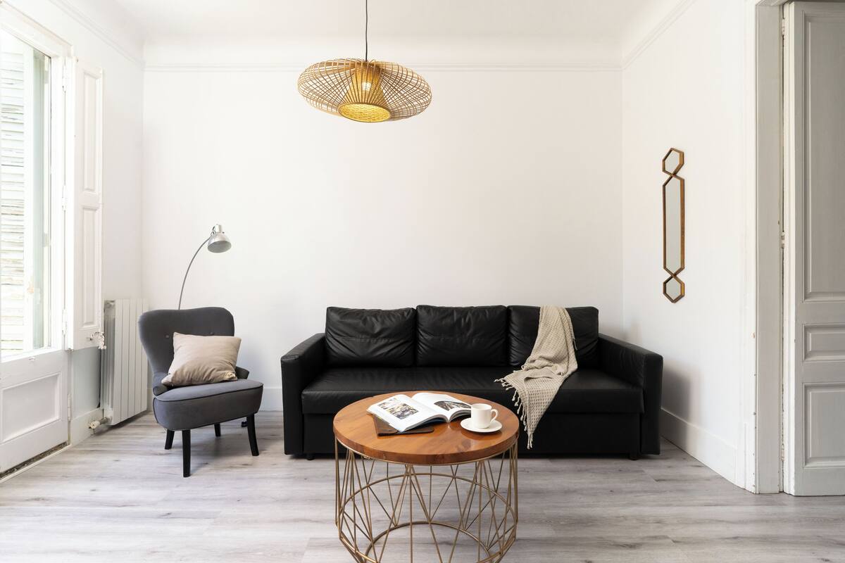 A modern living room with a black leather sofa, a round wooden coffee table, a grey armchair, and a pendant light.