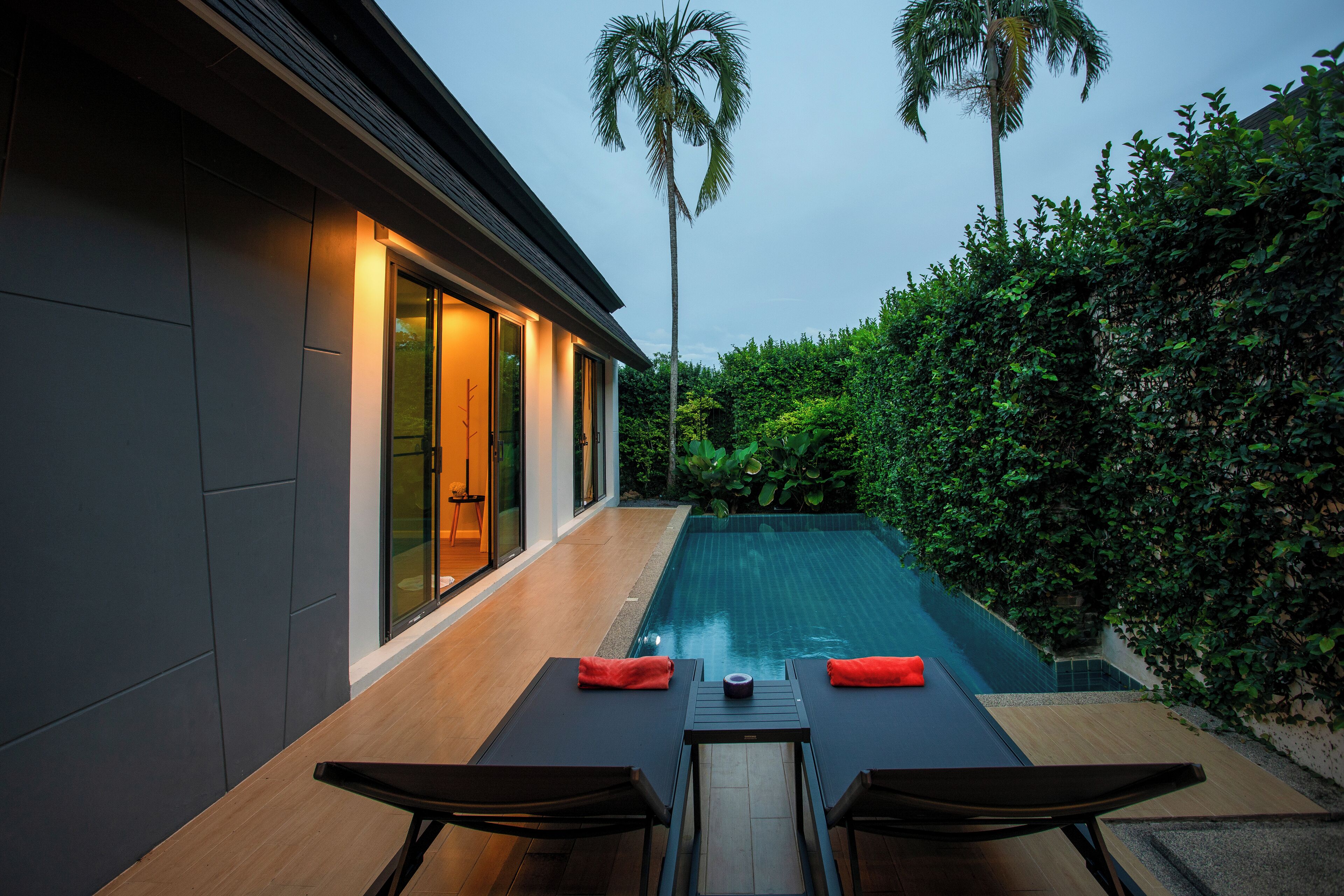 Una casa moderna con piscina, dos sillones de relax y una mesa con cojines rojos.
