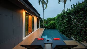 Una casa moderna con piscina, dos sillones de relax y una mesa con cojines rojos.