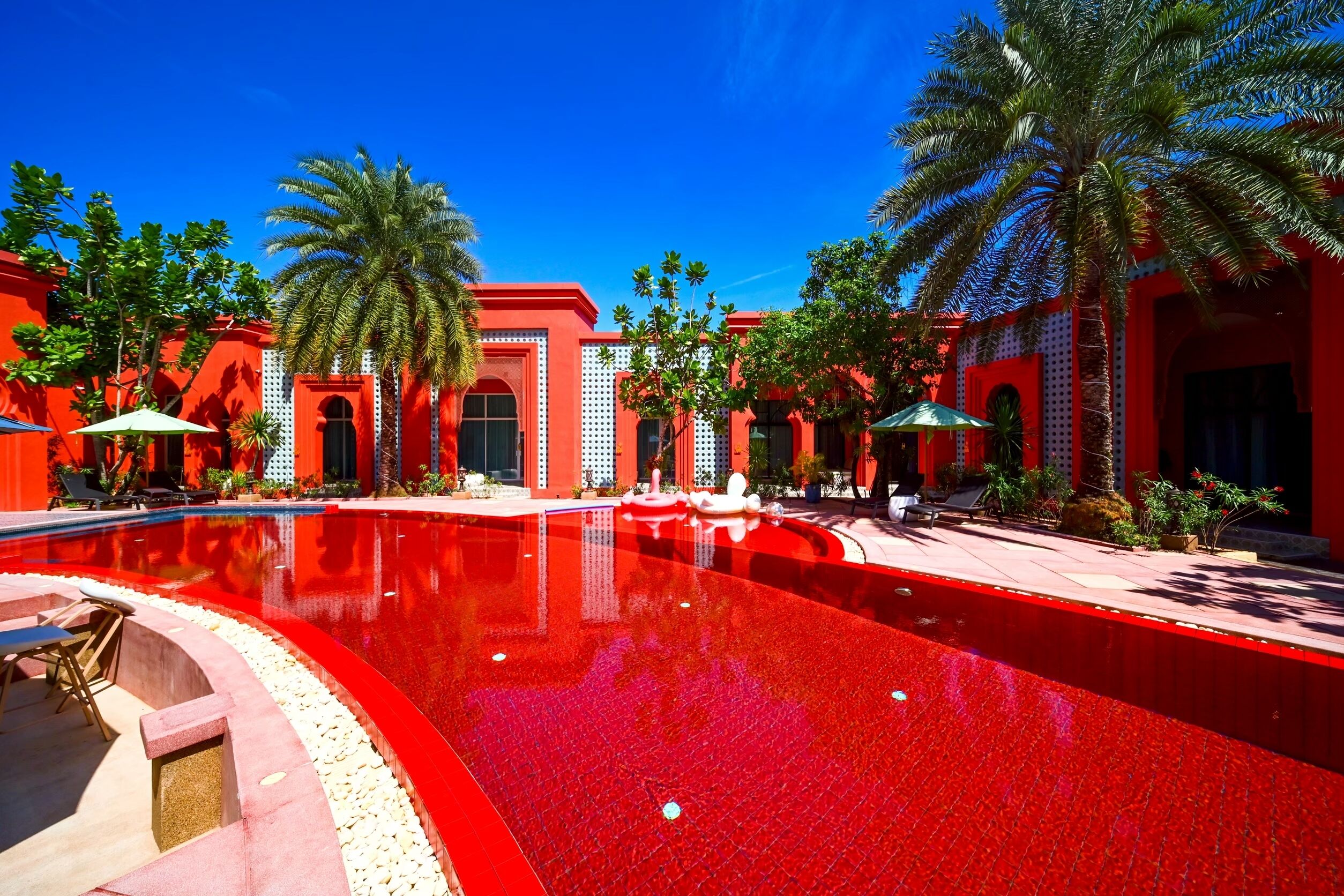 Un espace piscine doté d’un sol carrelé de tuiles rouges, de palmiers et de bâtiments rouges aux entrées en arc de cercle.