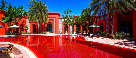 Un espace piscine doté d’un sol carrelé de tuiles rouges, de palmiers et de bâtiments rouges aux entrées en arc de cercle.