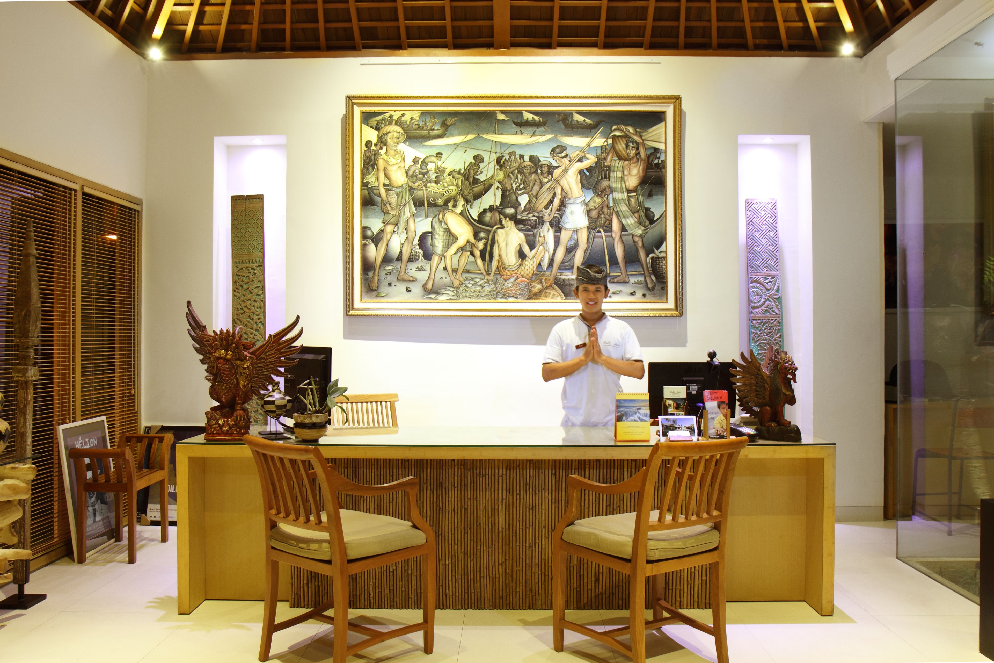 A reception area with a wooden reception desk, two wooden chairs, a large painting on the wall, and a person standing behind the desk.