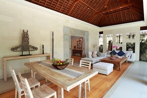 A spacious living area with a wooden dining table, white sofas, and a large window with a view of greenery. - Villa Bali Asri Batubelig (Seminyak)
