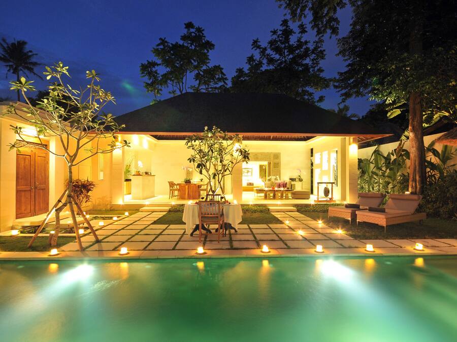 A poolside area with a dining setup and lounge chairs, surrounded by a well-lit patio and a swimming pool.