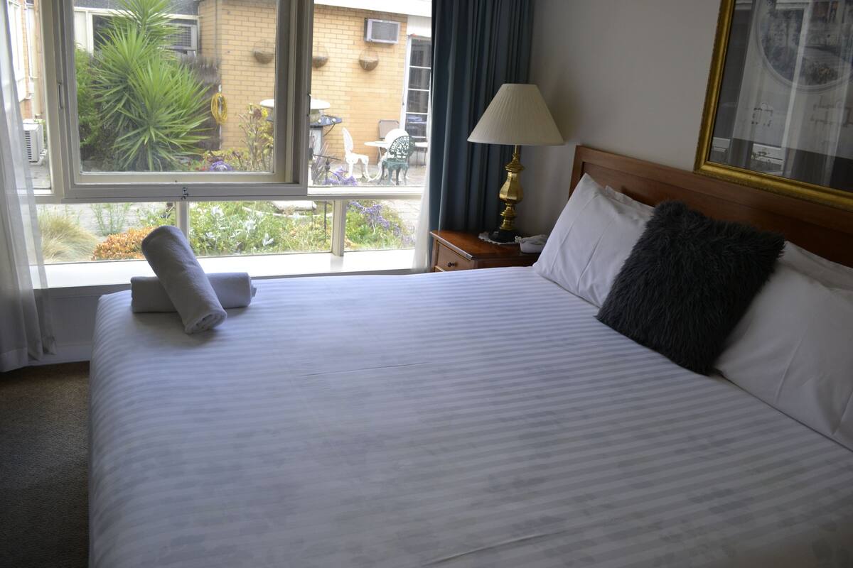 A hotel room with a large bed, a window with a view of the outside, a nightstand with a lamp, and a framed picture on the wall.