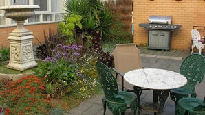 A patio area with a round table and four chairs, a barbecue grill, a stone urn, and various plants and flowers. - Aberdeen Motor Inn (Newtown)