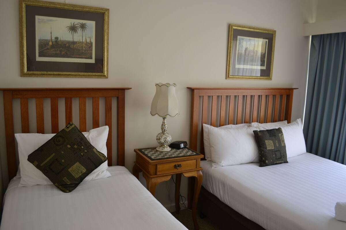 A hotel room with two single beds, a nightstand with a lamp, and two framed pictures on the wall.