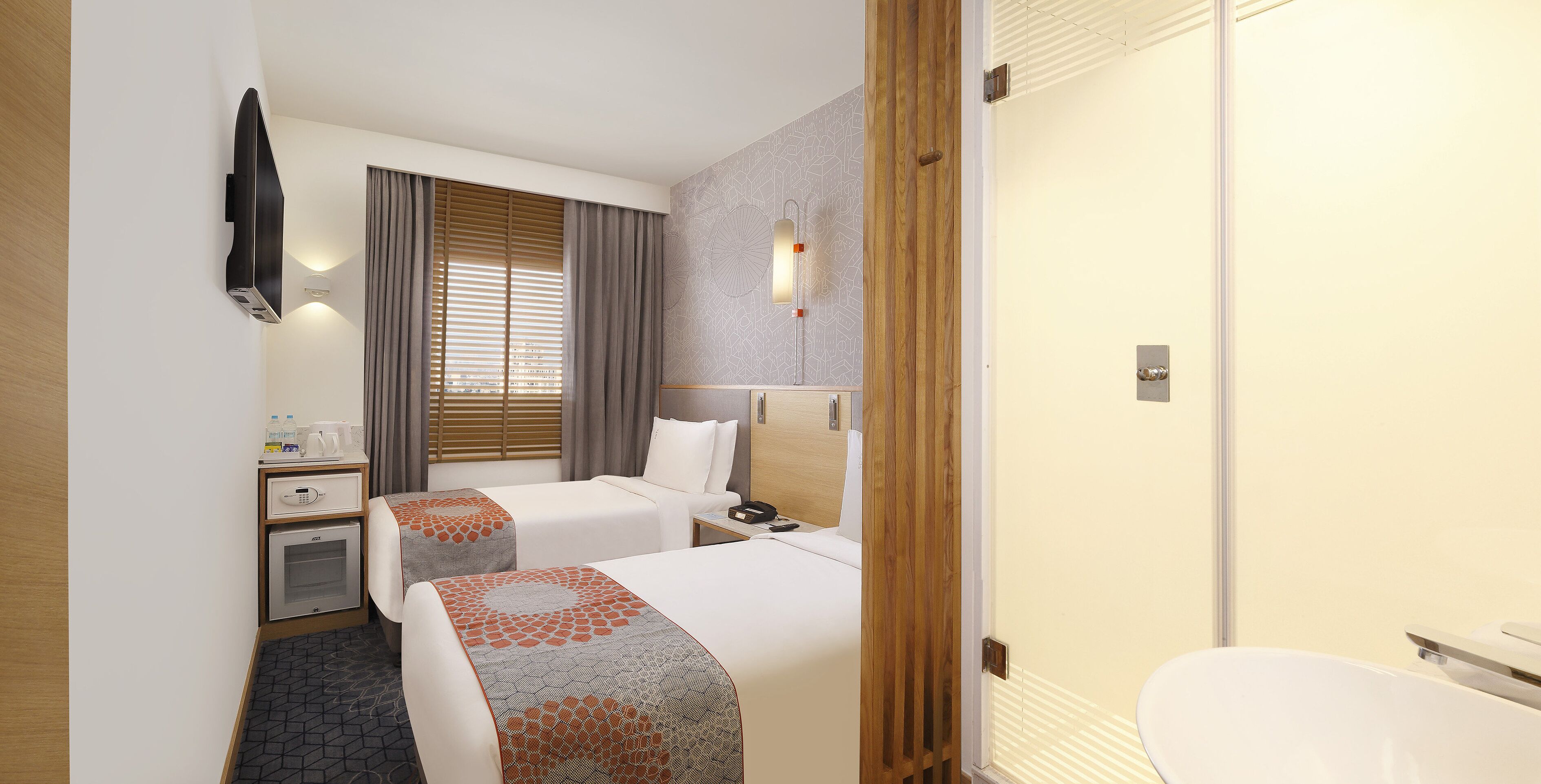 A hotel room with two beds, a desk with a telephone, a small table with a lamp, a shower area, and a bathroom.