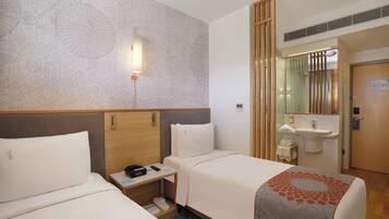 A hotel room with two beds, a bedside table with a phone and a lamp, a wall with a geometric pattern, a wooden door, and a bathroom with a sink and mirror.