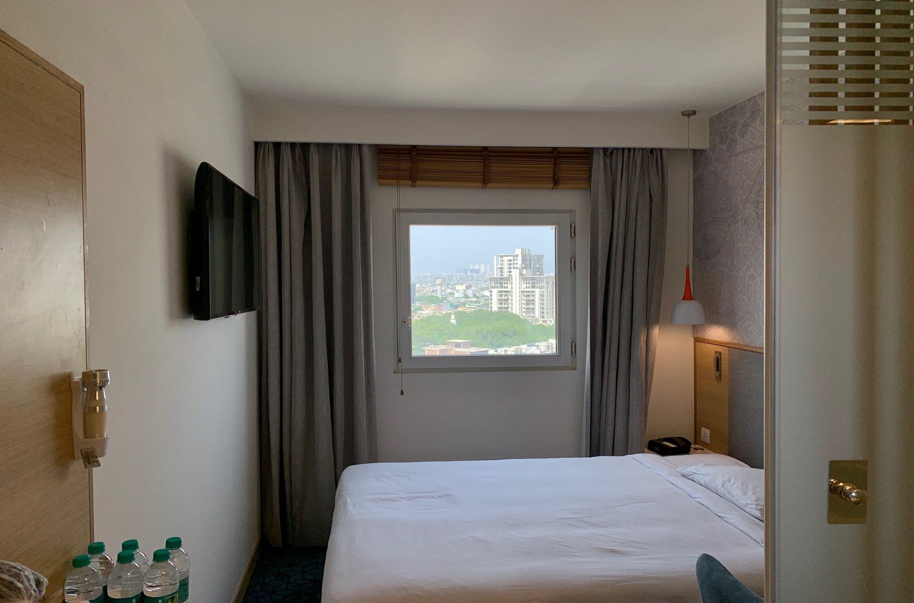 A hotel room with a bed, a window with curtains, a flat-screen TV mounted on the wall, and a bedside table with bottled water.