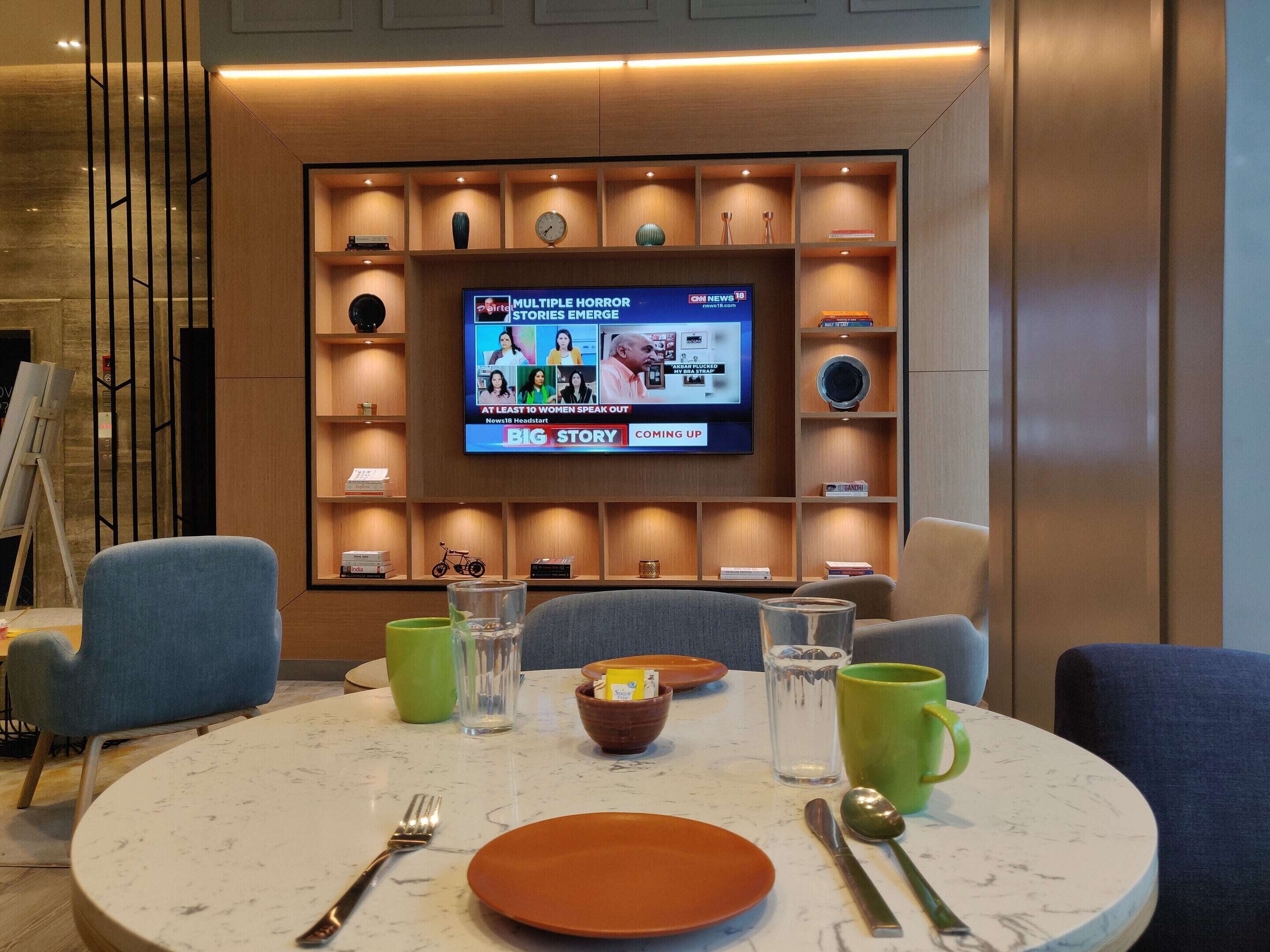 Quarto de hotel moderno com uma mesa de jantar redonda, cadeiras azuis e uma televisão exibindo um programa de notícias.