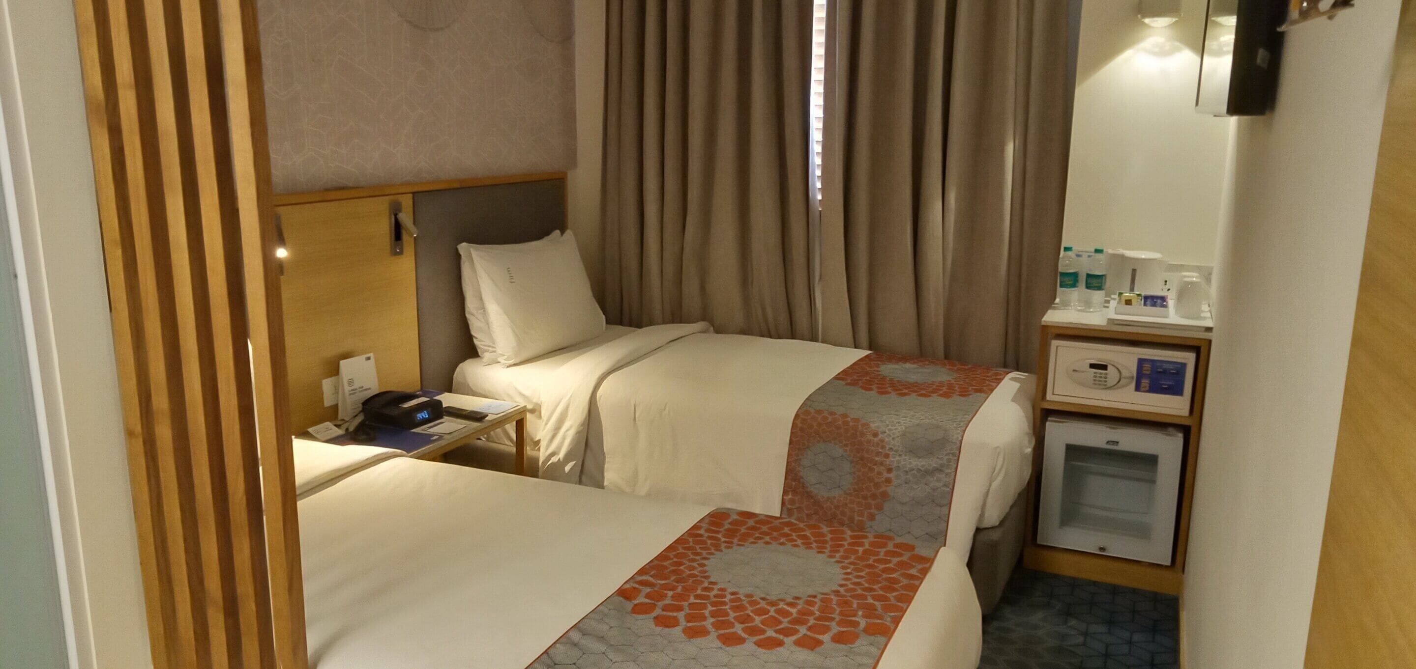 A hotel room with two beds, a bedside table with a phone, a small desk with a computer, a microwave, and a mini-fridge.