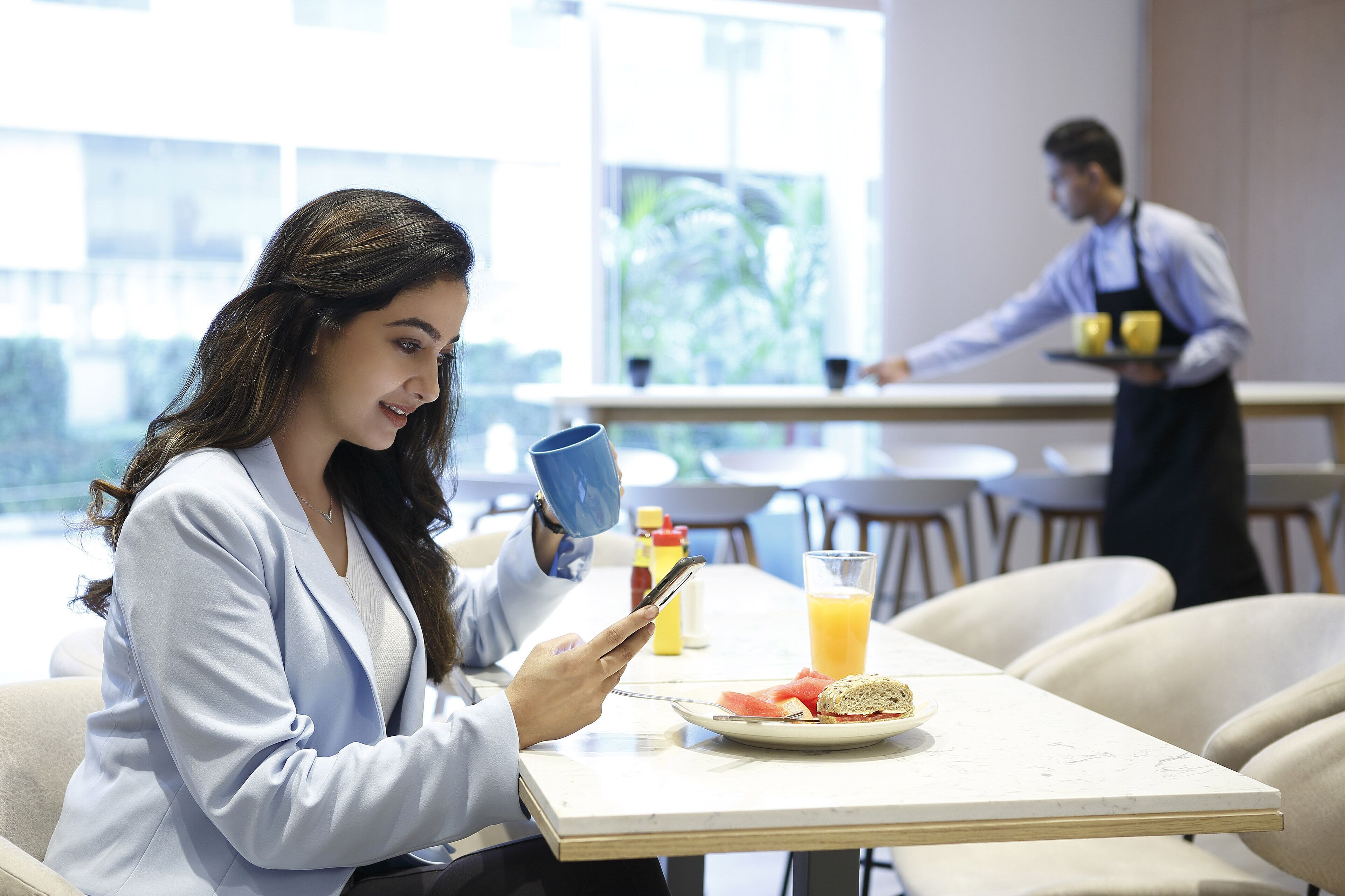 A person using a smartphone at a table with a plate of food and a drink, in a bright room with large windows.