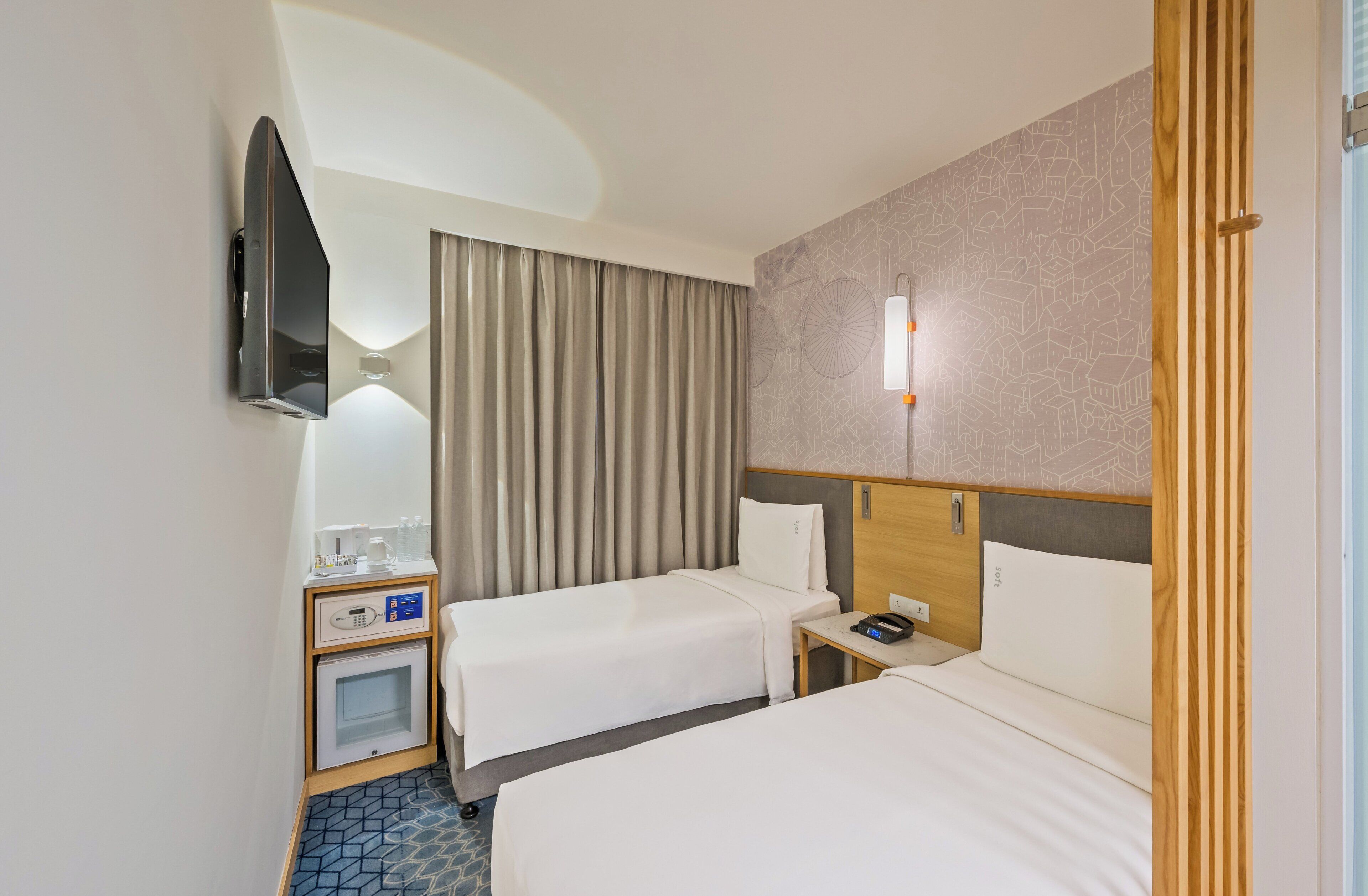 Camera d'albergo con due letti, una TV a schermo piatto montata a parete, un minibar, un tavolino con telefono e lampada, e una parete decorata.