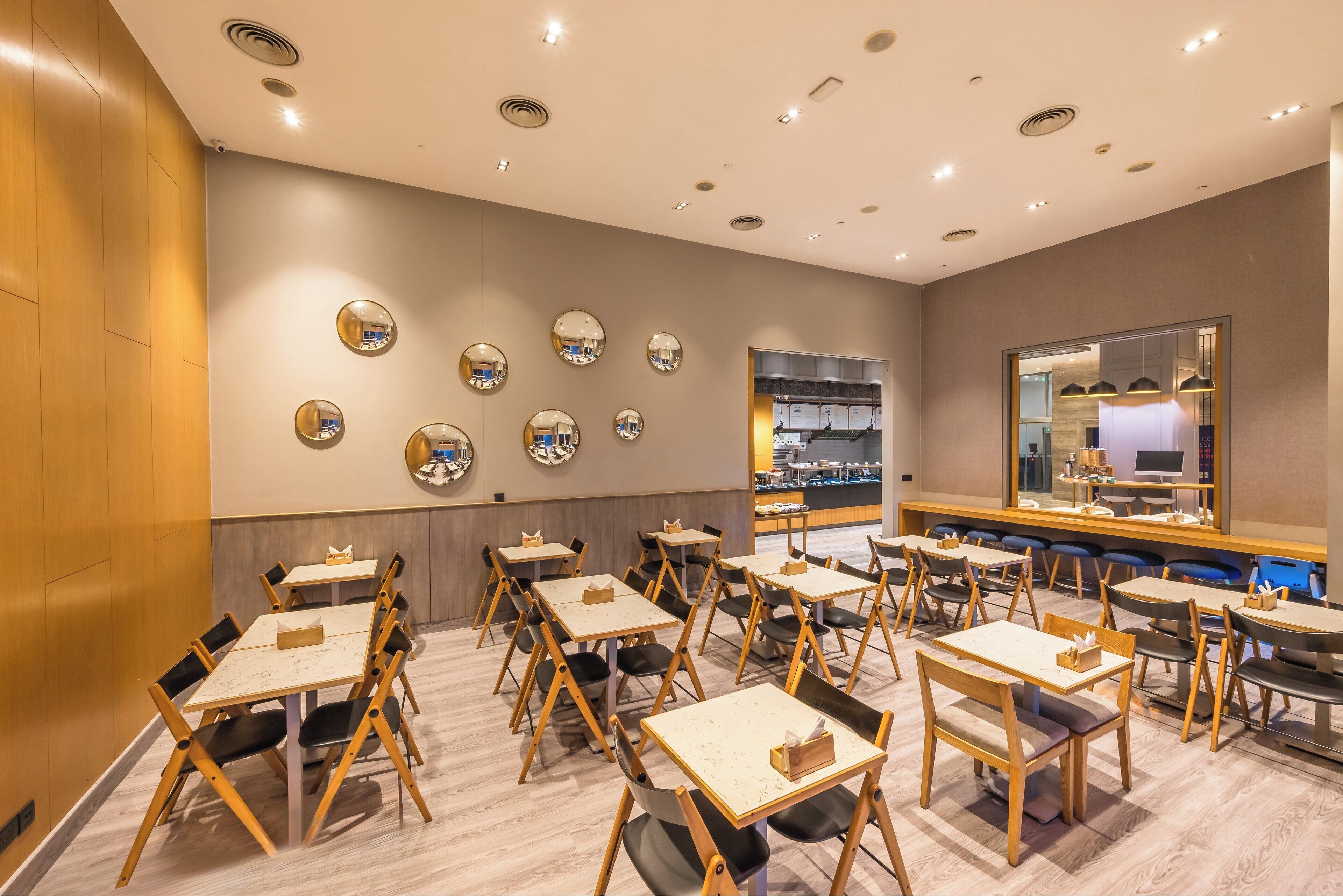 A modern dining area with multiple tables and chairs, a wall with circular mirrors, and a view of the kitchen through a glass partition.