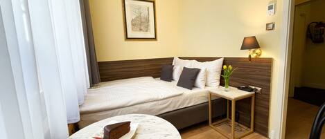 A hotel room with a bed, a small table with a cake, a vase with flowers, a lamp, and a framed picture on the wall.