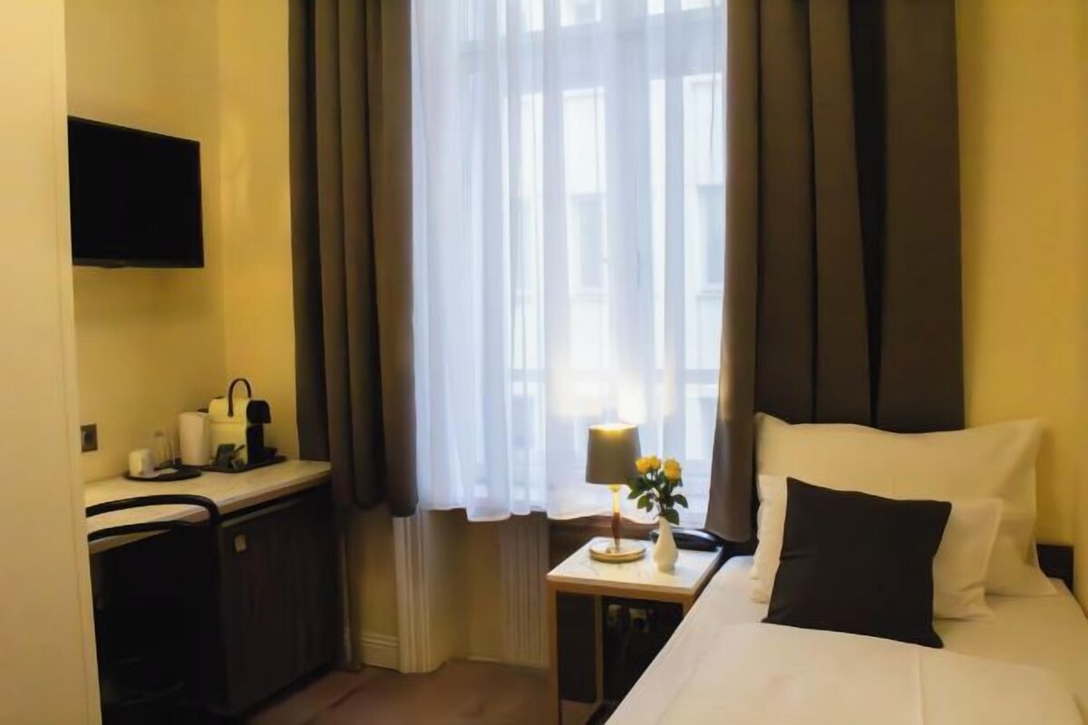 A hotel room with a bed, a small bedside table with a lamp and vase, a TV mounted on the wall, and a window with curtains.