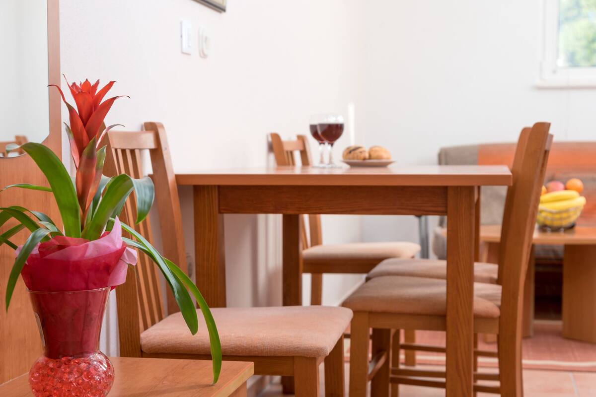 Ein Essbereich mit Holzsesseln und einem Tisch, einer Vase mit einer roten Blume sowie einem Teller mit Brot und einem Glas Rotwein.