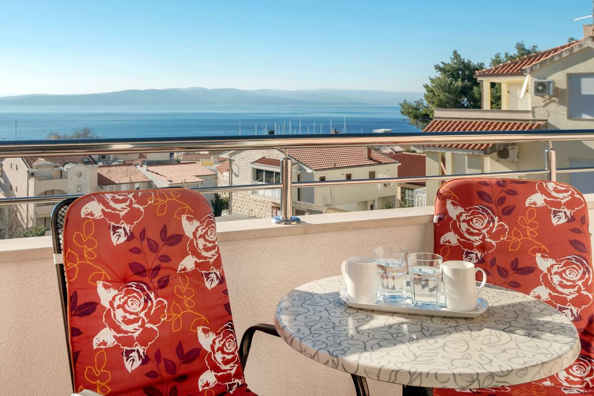 Ein Balkon mit Tisch und Stühlen, der Blick auf die Küste mit Gebäuden und dem Meer freigibt.