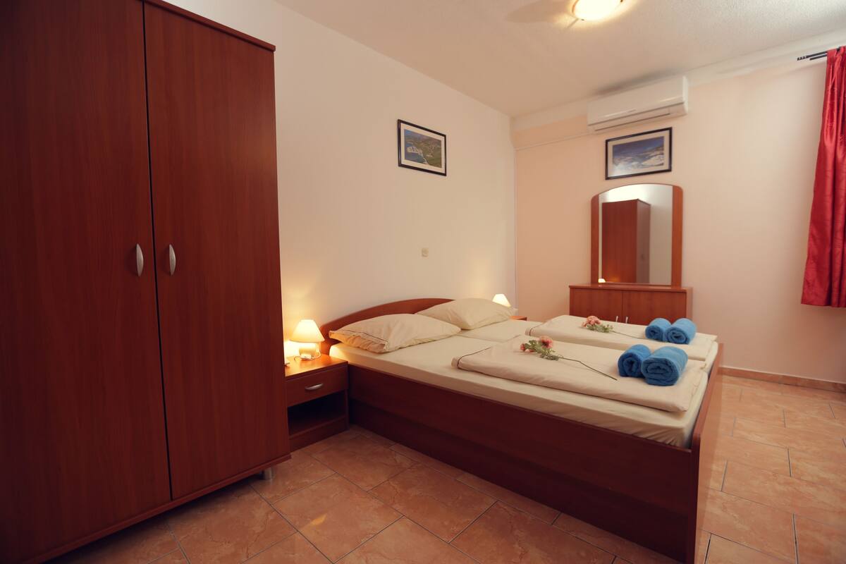 Ein Schlafzimmer mit einem Holfischschrank, einem Bett mit Kissen und Handtüchern, einem Nachttisch mit Lampe und einem Wandspiegel.